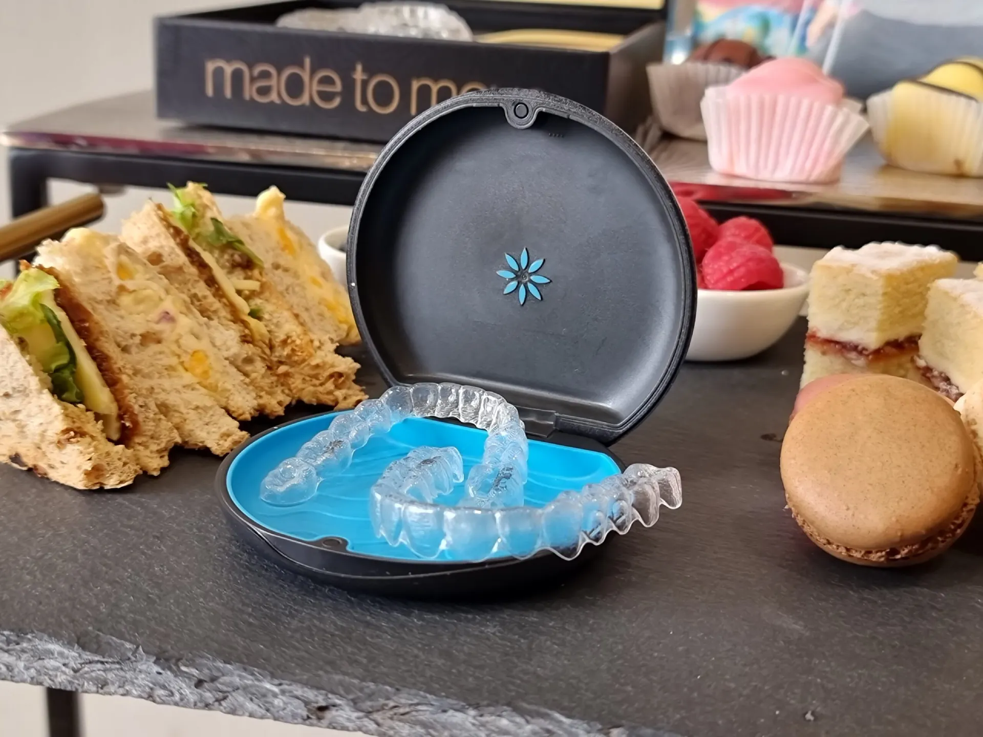 Clear aligners in a blue-lined case beside sandwiches and desserts on slate tray.